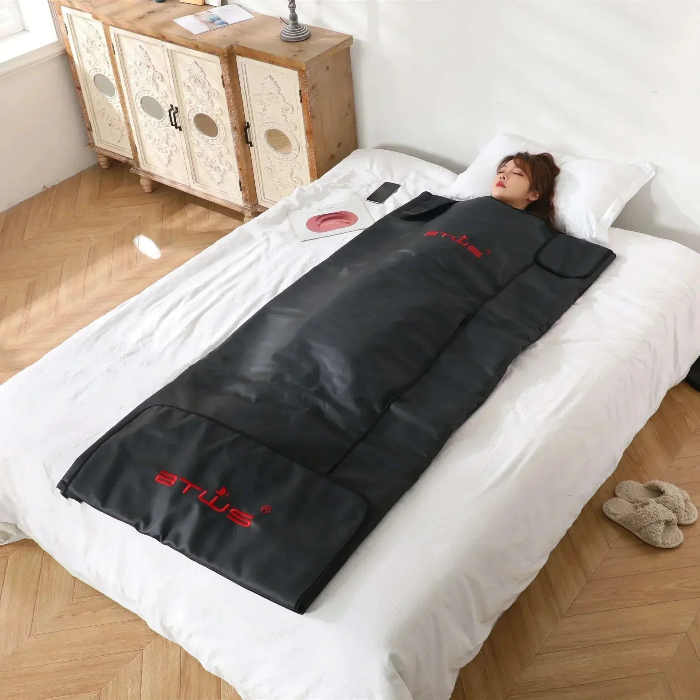 Person relaxing in a black infrared sauna blanket on a white bed in a bright room for weight loss and detox therapy